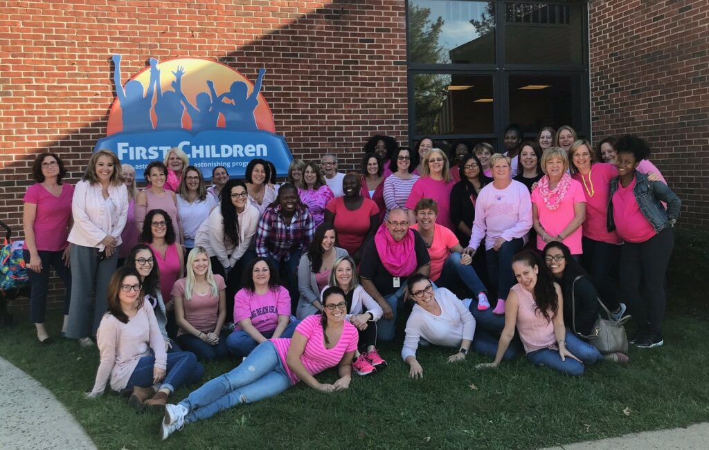 A group of teachers huddled together smiling for a photo. They are outside and wearing pink shirts.