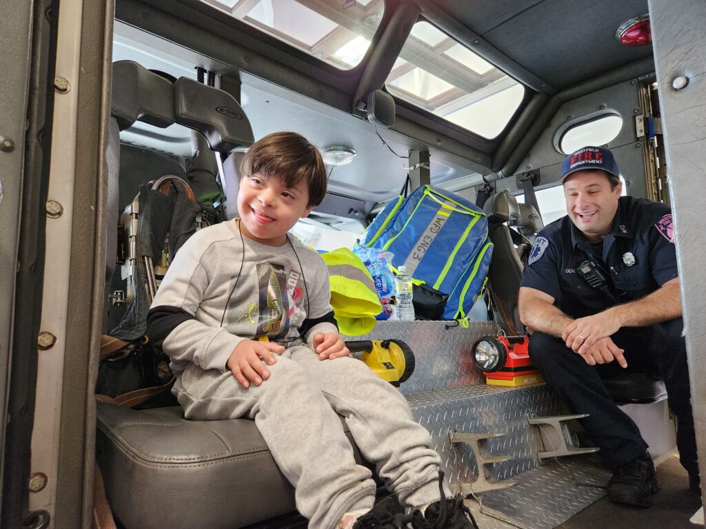 A light skinned child sit with a light skinned male adult in a fire truck. They are smiling.