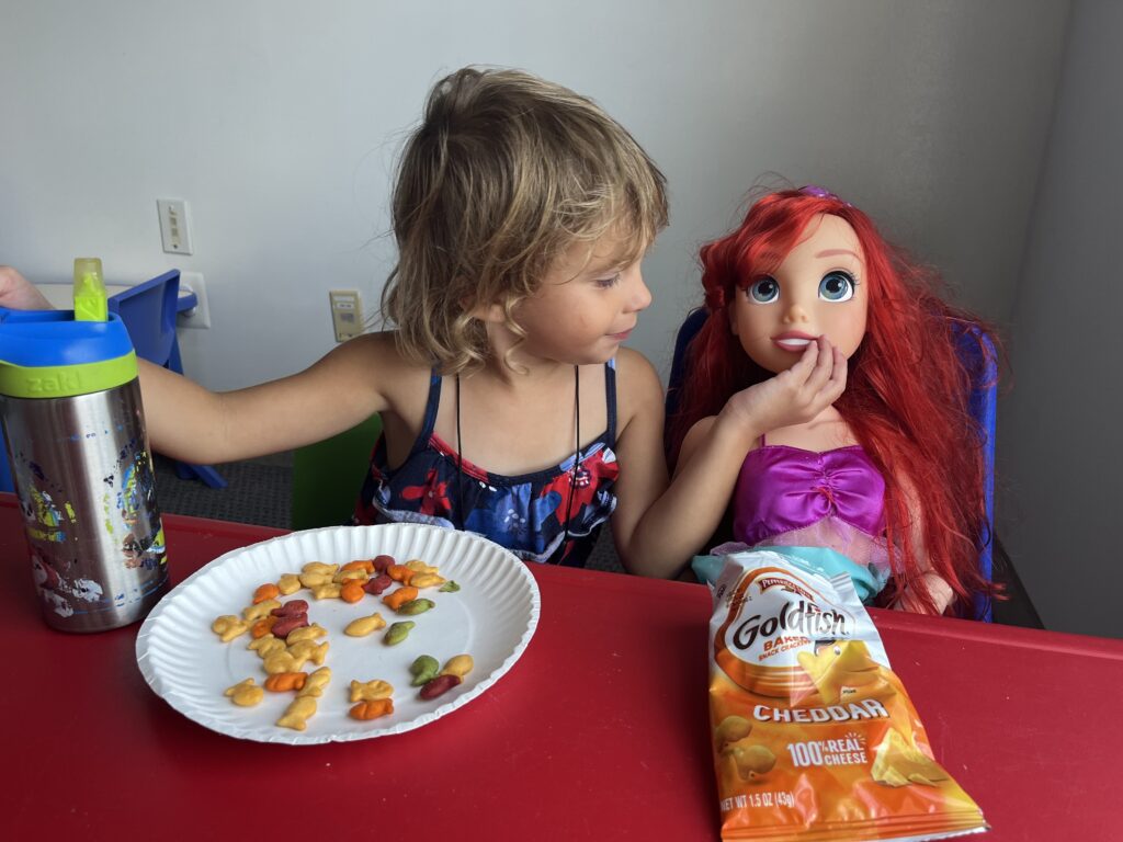 A young girl plays with her Disney princess doll at a table. They're eating goldfish crackers.