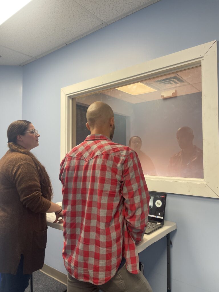A light skinned female and male stand at a two way mirror. They are looking into a clinical sesson.
