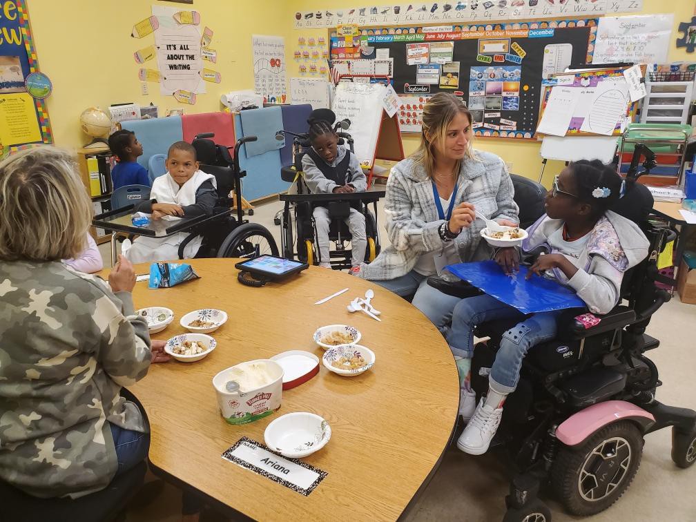 A classroom with students. There are wheelchairs and a table, with a teacher.