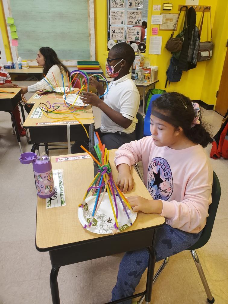 Students in their classroom, one student is sitting at her desk and she is playing with a model she made.